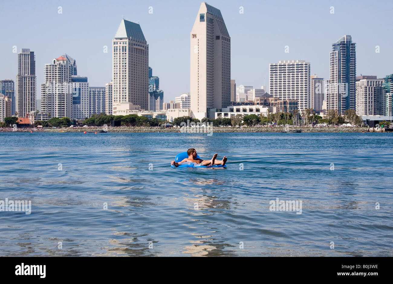 A man floats on a small blue raft, with the San Diego skyline in the background.  It is a beautiful summer day. Stock Photo
