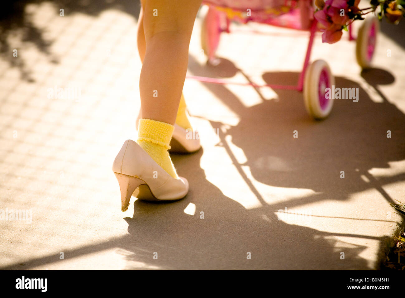 young girl walking in mom's shoes, pushing toy baby stroller, color, close up shoes and leg Stock Photo