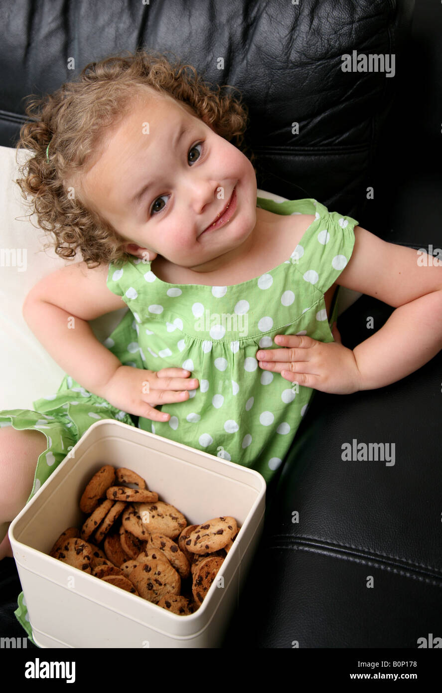 A cheeky 3 year old girl sitting on the sofa at home with a tin of cookies on her lap putting her hands on her hips Stock Photo