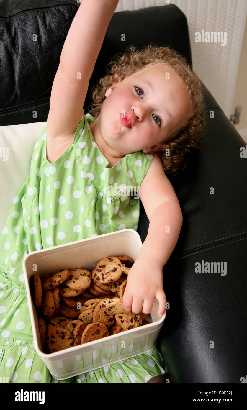 A naughty 3 year old girl sitting on the sofa at home eating biscuits Stock Photo