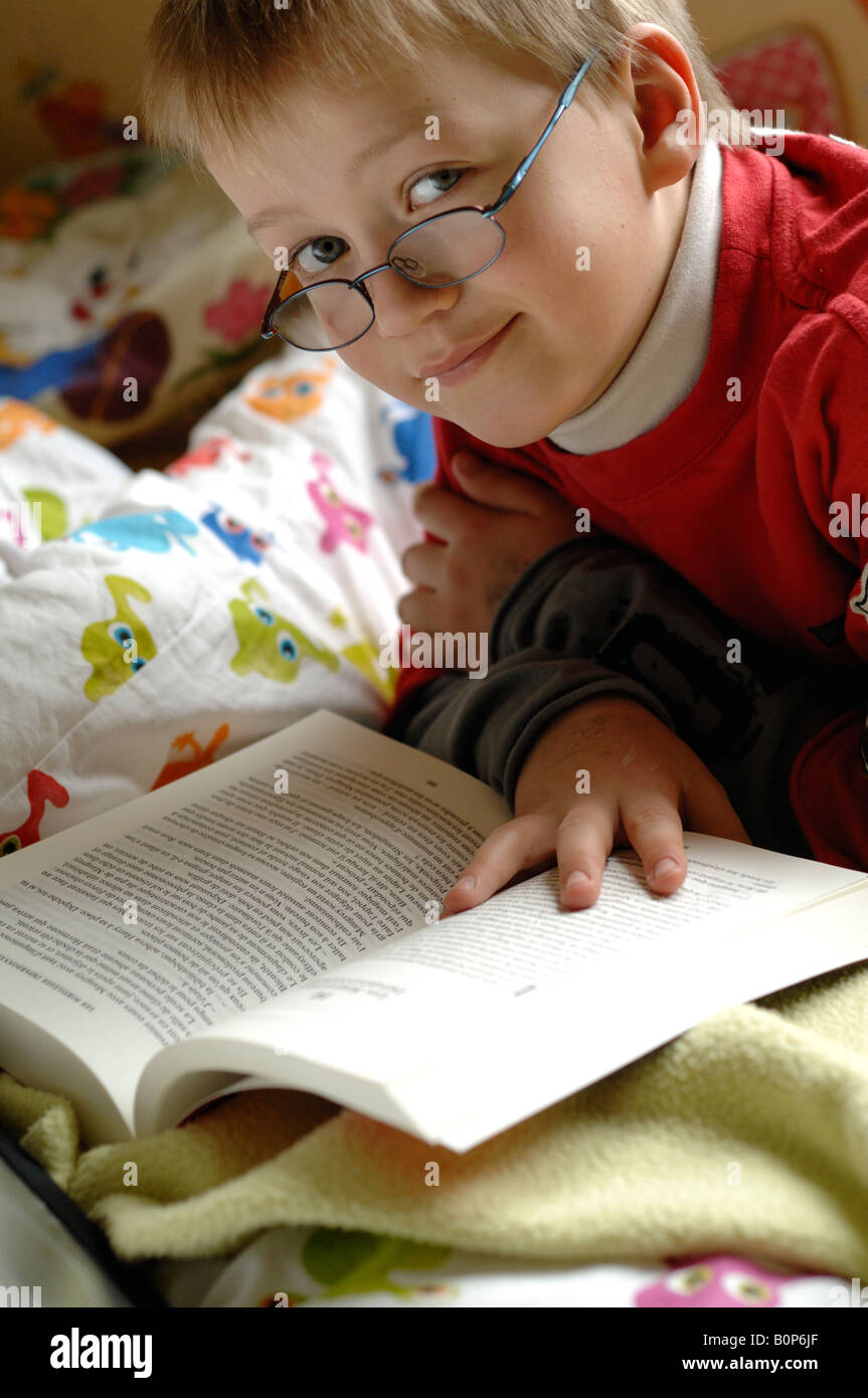LITTLE BOY READING A BOOK, WEARING GLASSES, on his bed Stock Photo