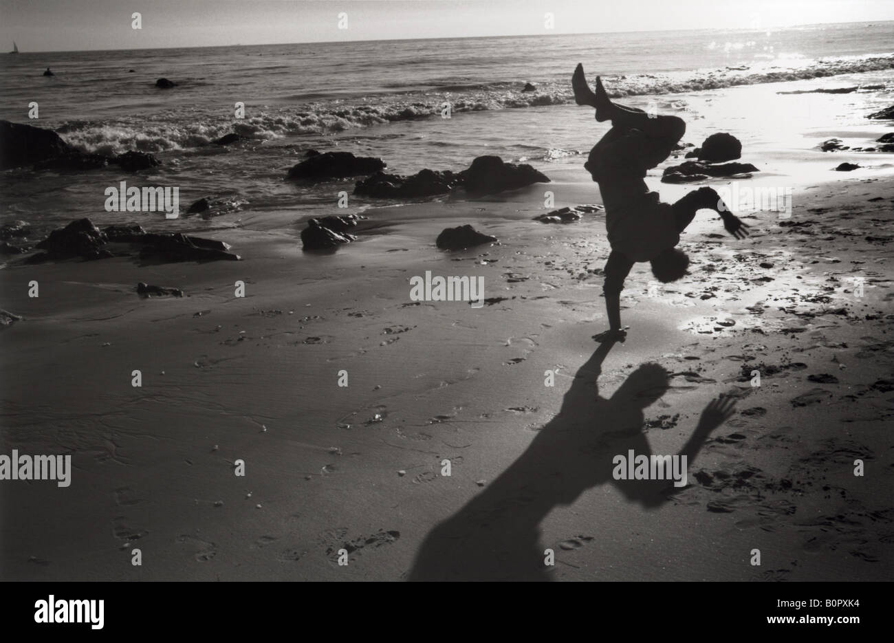 silhouette of man doing one handed cartwheel on beach shore on sand black and white horizontal Stock Photo