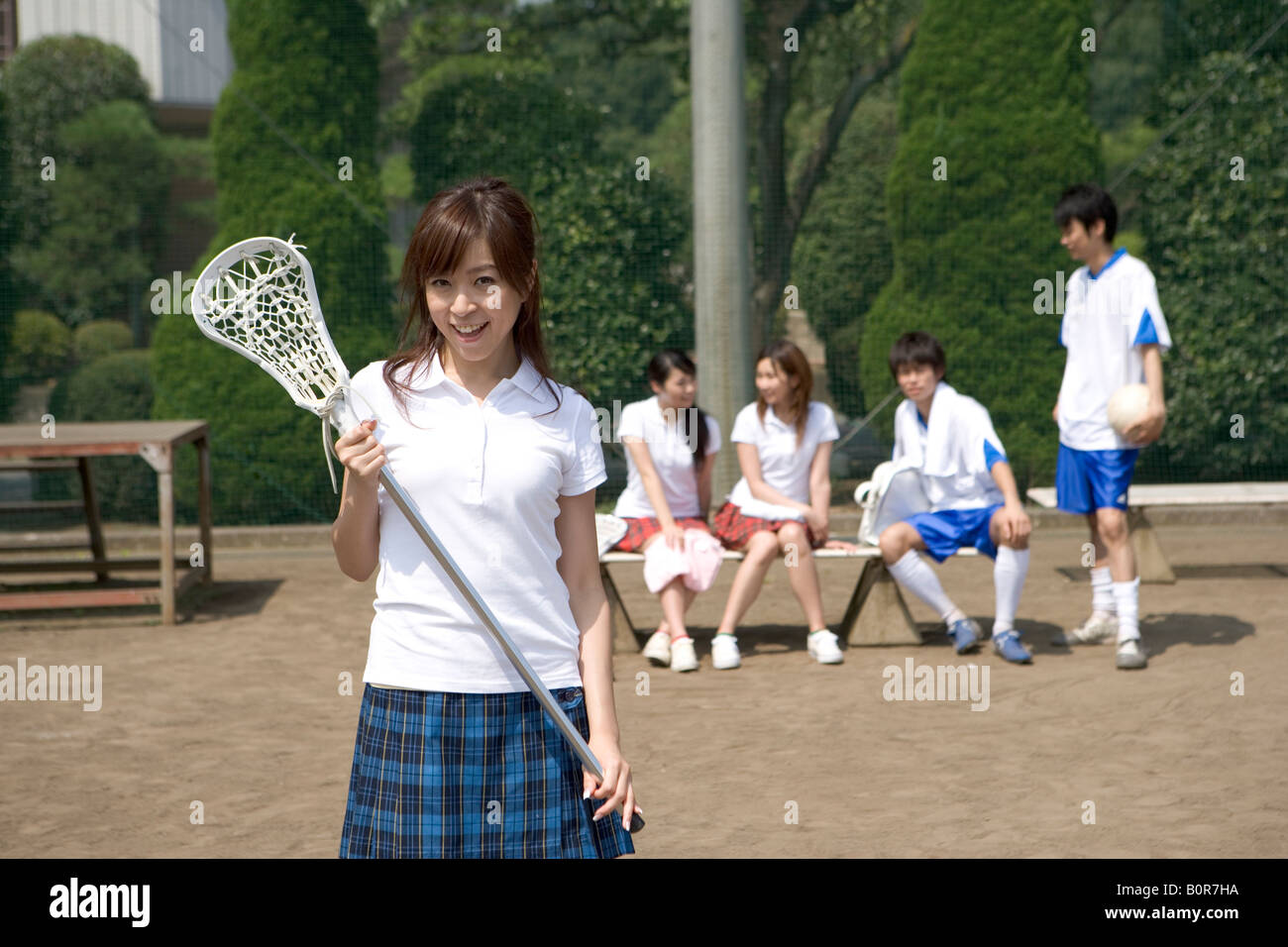 Teenage girl standing and holding lacrosse stick with friends sitting on bench in background, portrait Stock Photo