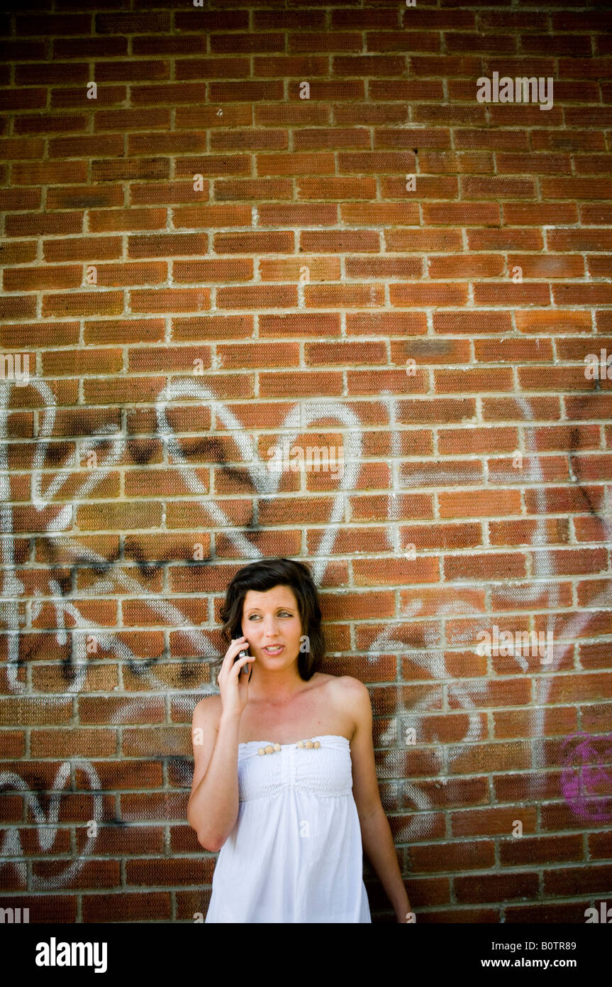 Young teenage woman talking on her mobile phone leaning against a graffiti covered brick wall.UK Stock Photo