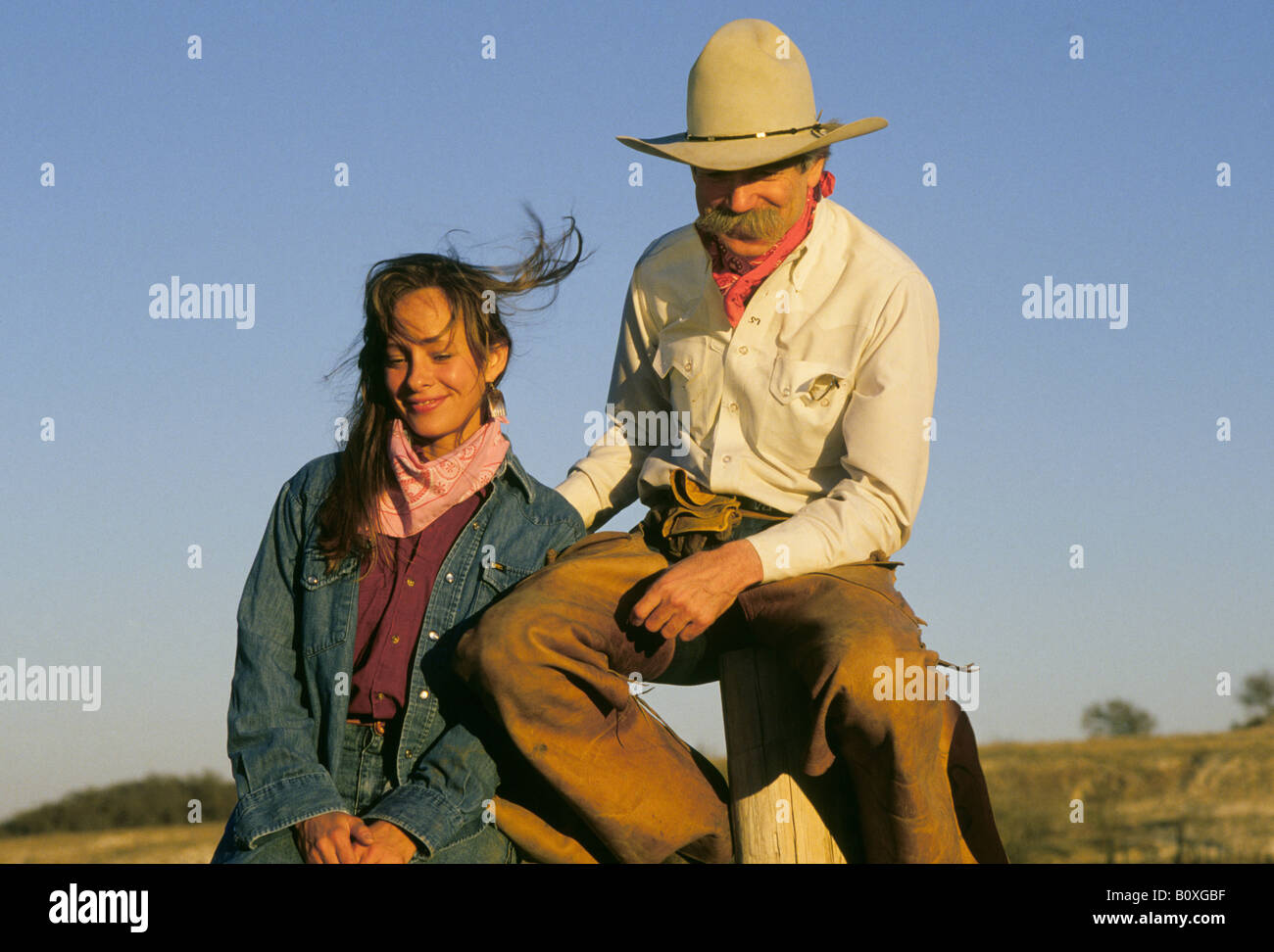 A cowgirl guest at a dude ranch near San Antonio Texas sits on a fence with a cowboy wranger in chaps and a big hat at sunset Stock Photo
