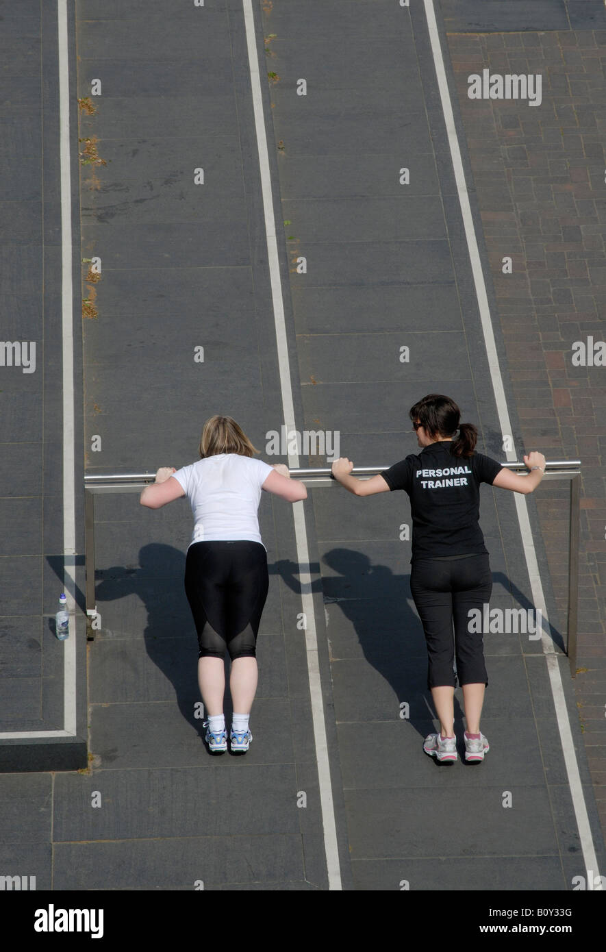 Personal trainer (indicated on the back of her t-shirt) and client doing push ups on raised bar, South Bank, London, England Stock Photo