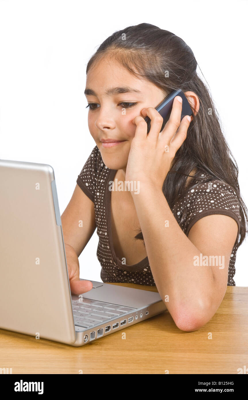 A young girl (10 yrs) at a desk with a lap top computer while talking on a mobile phone against a pure white (255) background. Stock Photo