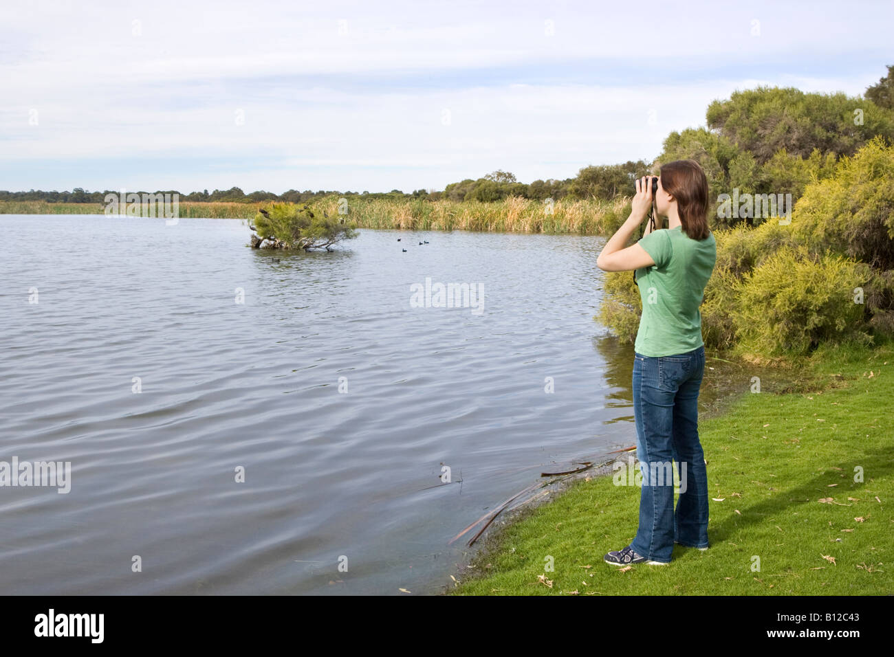 A mid 20s woman with binoculars birdwatching at the side of a lake ...