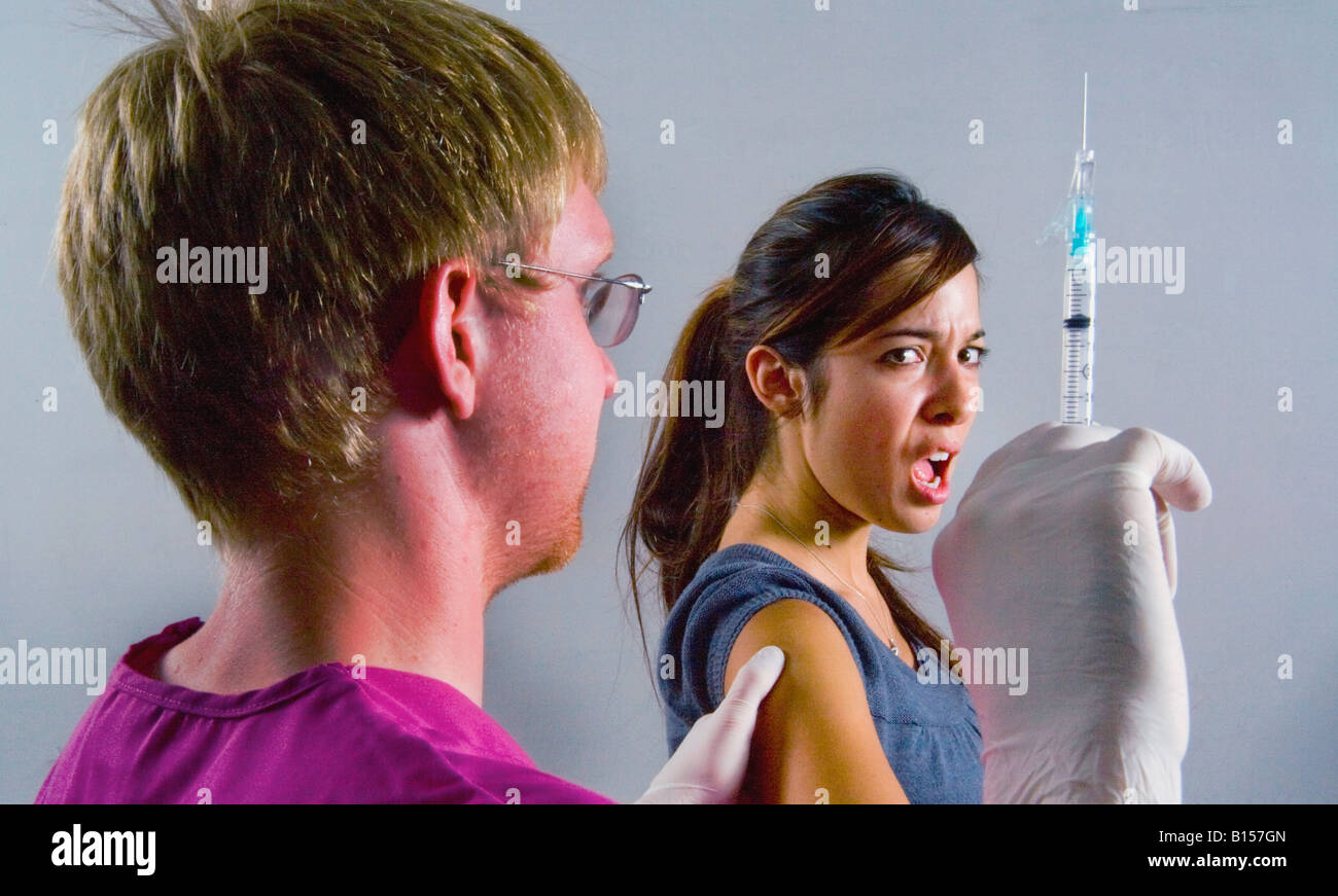 A young woman winces at the prospect of an injection as a medical professional brandishes a hypodermic syringe Stock Photo