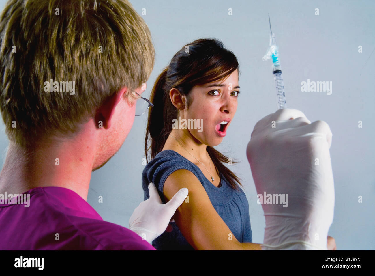 A young woman winces at the prospect of an injection as a medical professional brandishes a hypodermic syringe Stock Photo