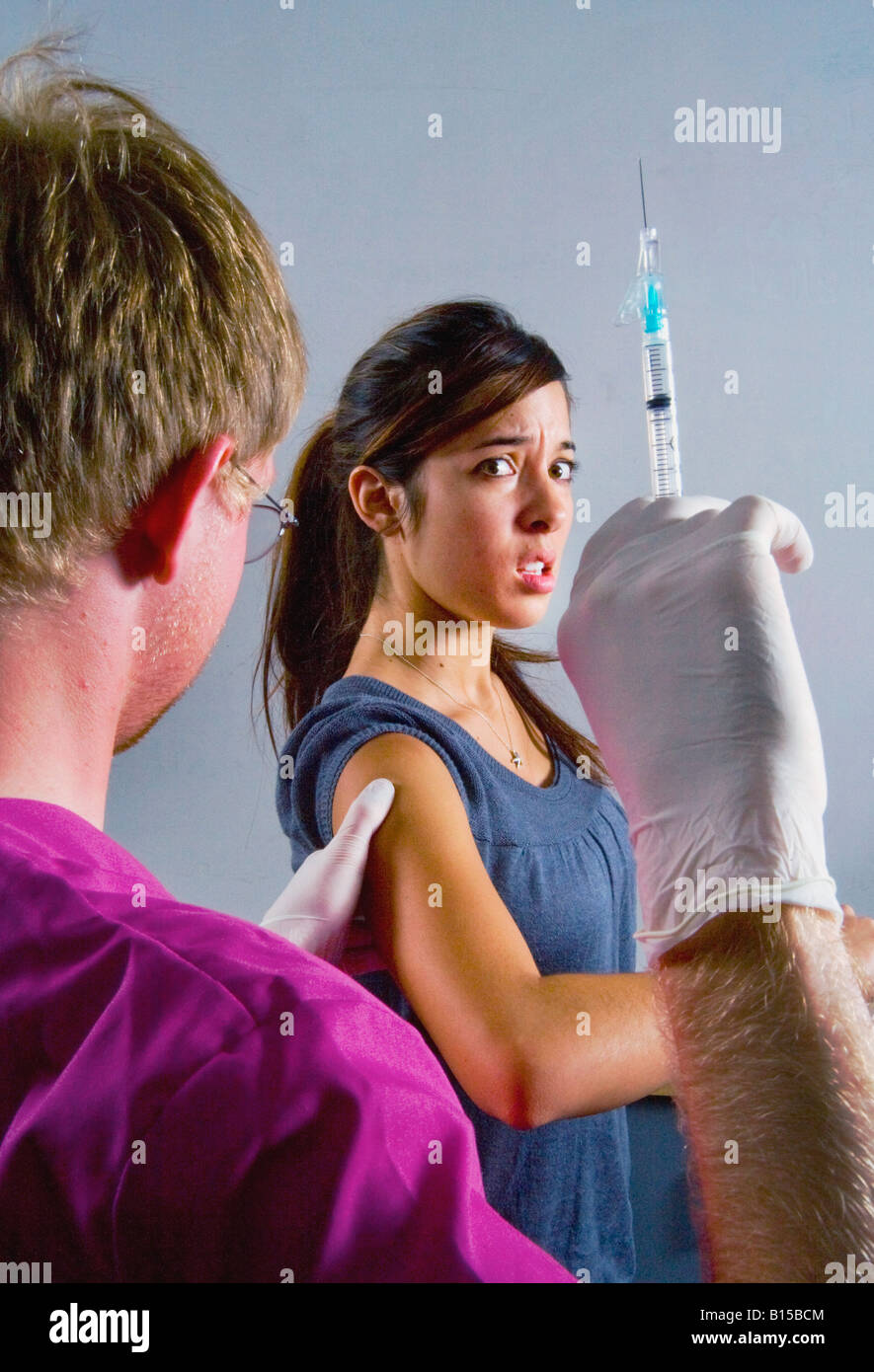 A young woman winces at the prospect of an injection as a medical professional brandishes a hypodermic syringe Stock Photo