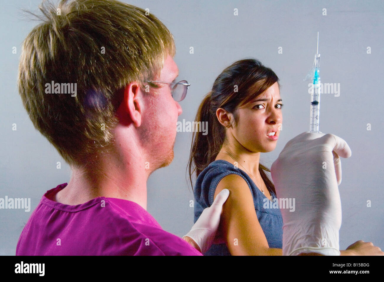 A young woman winces at the prospect of an injection as a medical professional brandishes a hypodermic syringe Stock Photo
