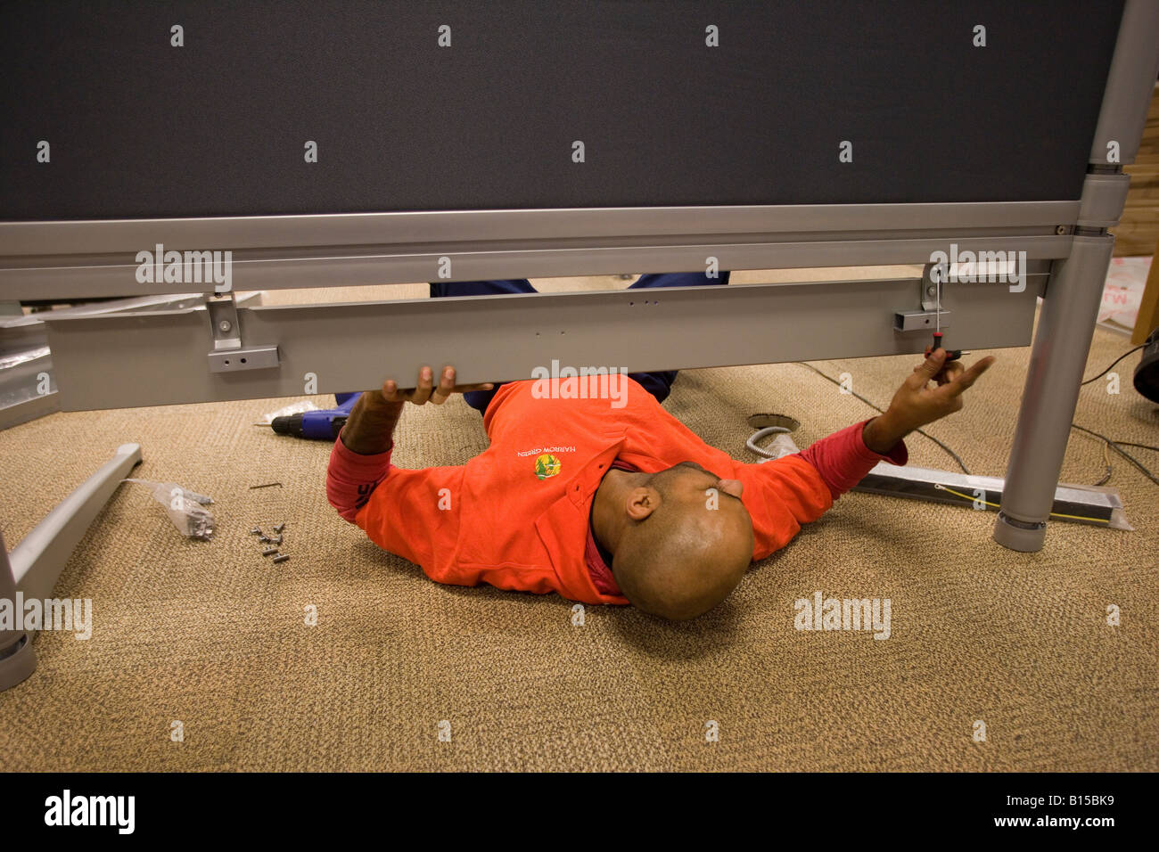 A workman lies on the floor to secure a desk fitting in a newly constructed office building. Stock Photo