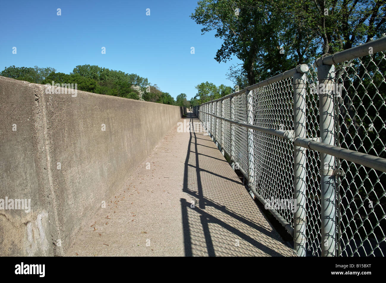 The vanishing points of a sidewalk it s concrete traffic barrier and chain link fence form a long straight linear perspective Stock Photo