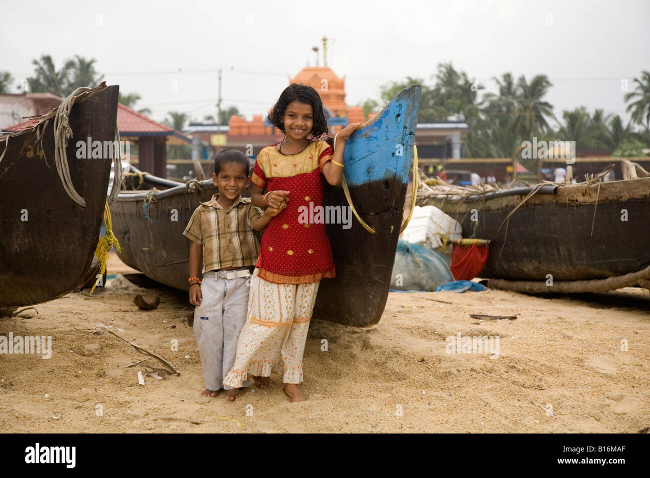 Two children grin for the camera on Kappil Beach in Kasaragod, Kerala. Fishing boats are pulled up on the beach behind them. Stock Photo