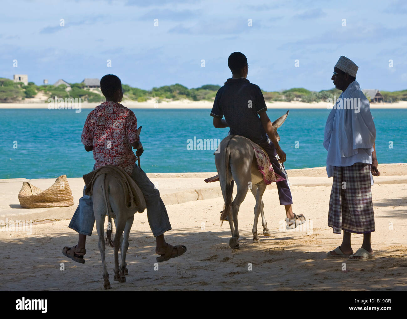 Kenya, Lamu Island, Shela. Two young men on donkeys ride past a Muslim man at Shela pier. Stock Photo