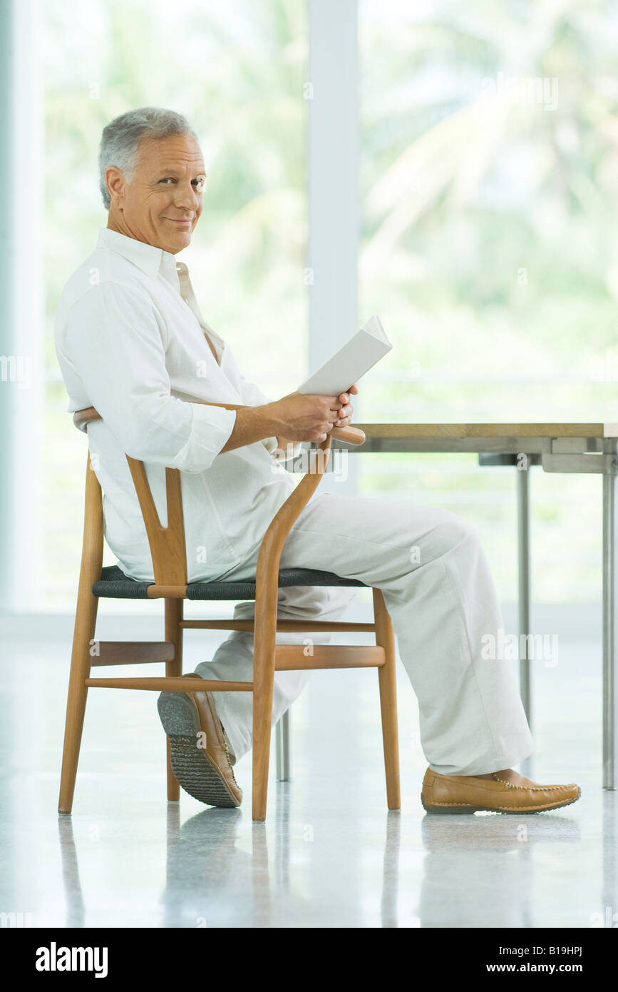 Mature man sitting at table, holding book, smiling at camera Stock Photo