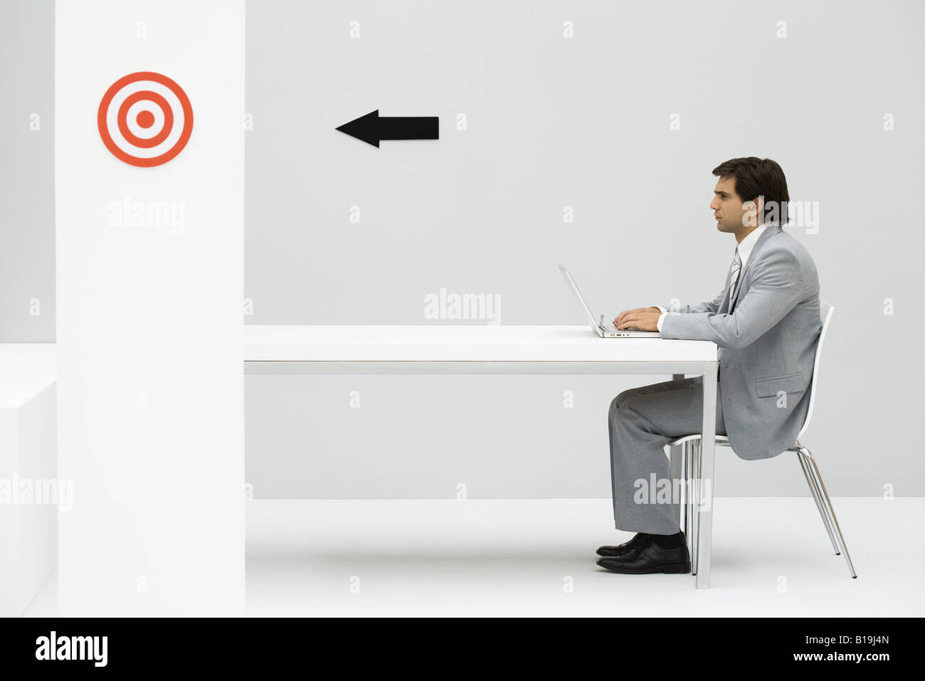 Man sitting at desk, using laptop computer, arrow on wall pointing at bull's-eye Stock Photo