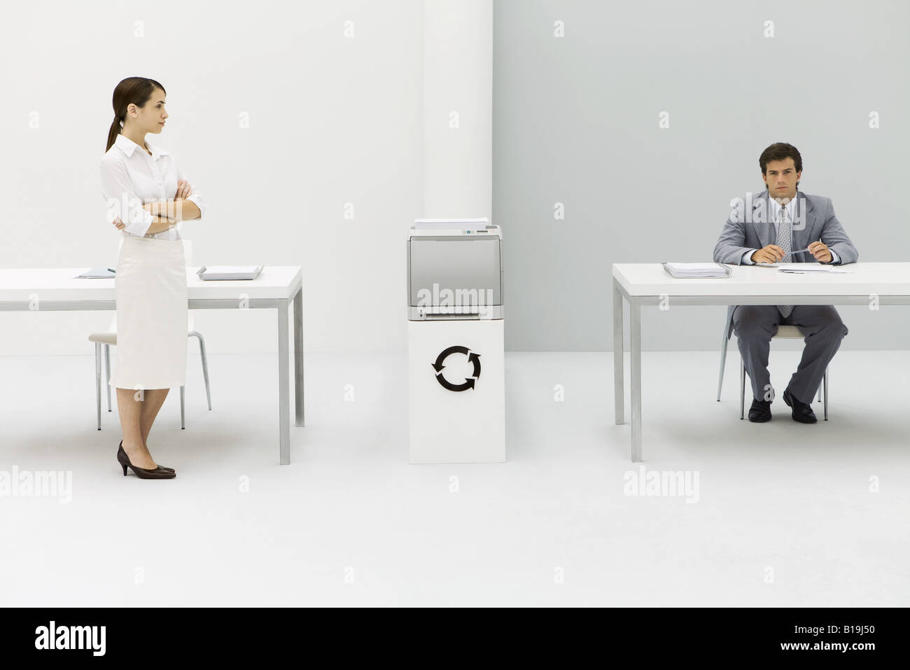 Two professionals in office, woman with arms folded, man looking at camera, circular arrow symbol between them Stock Photo