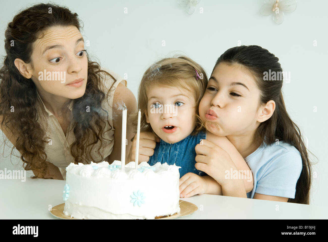 Girl helping younger sister blow out candles on birthday cake, mother watching Stock Photo