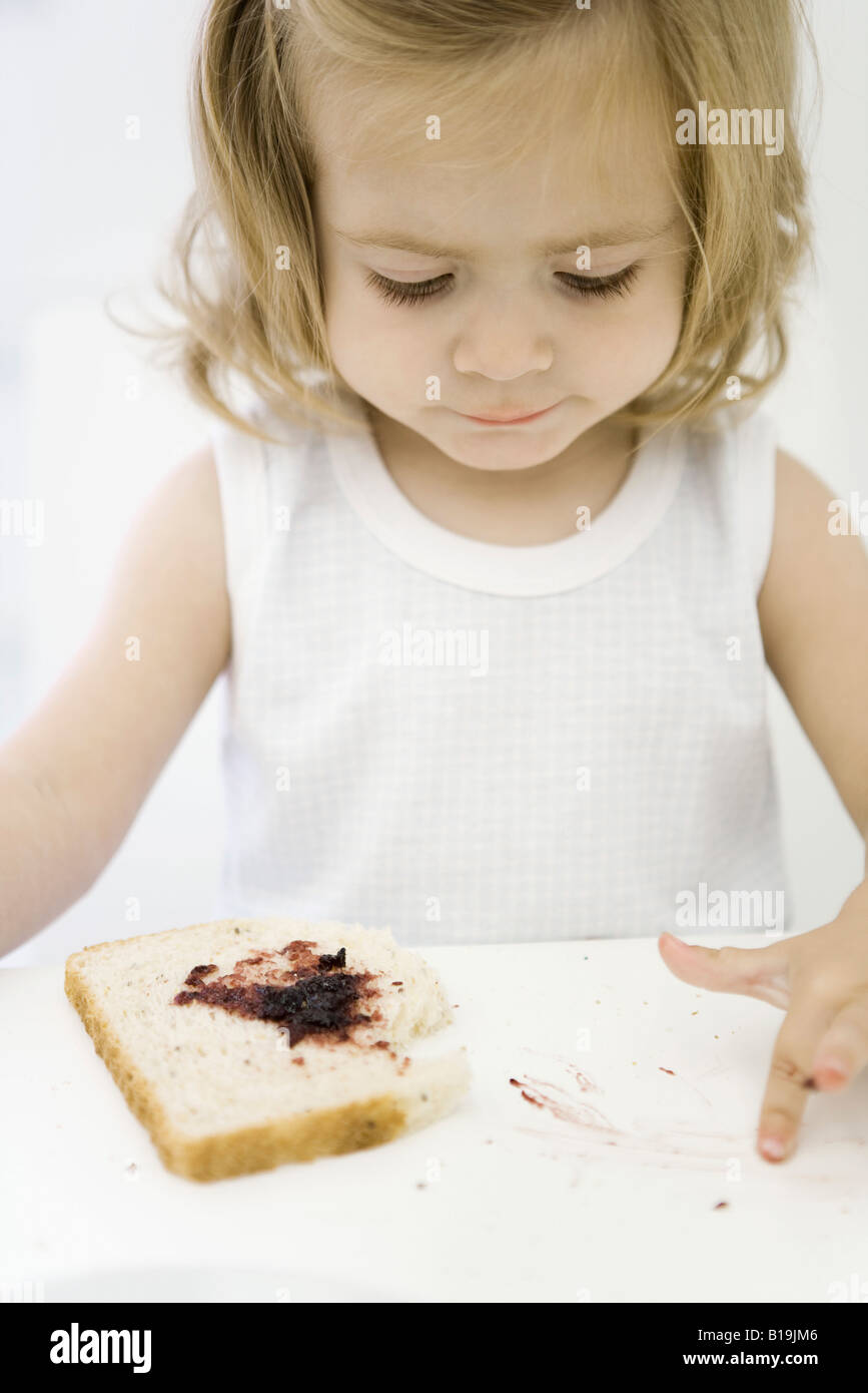 Little girl with slice of bread, spreading jam on table with finger