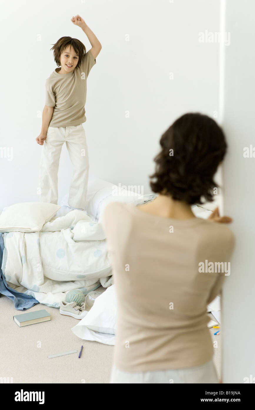Little boy jumping on unmade bed, mother watching from doorway Stock Photo