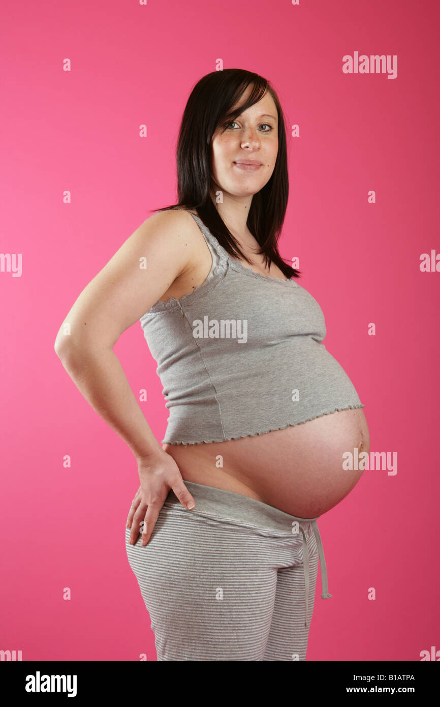 A side on view of a dark haired 38 week (nine months) pregnant woman standing hand on hips wearing casual grey clothes. Stock Photo