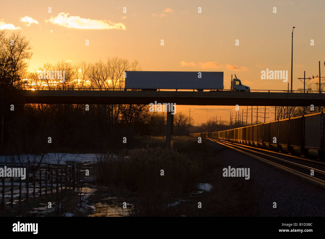 A tractor trailer truck passes over a loaded coal train as the sun sets. Stock Photo
