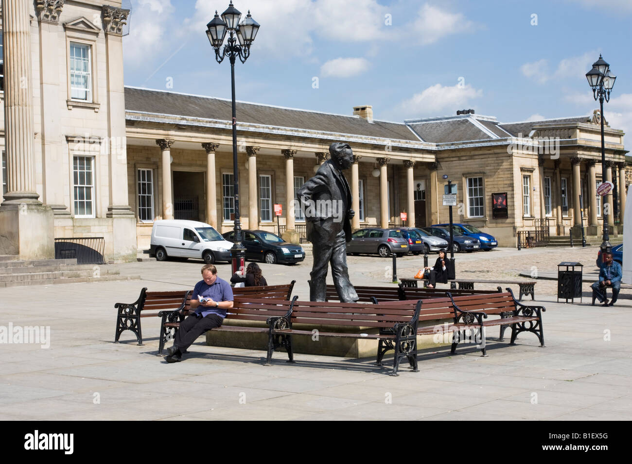 A man Sitting next to a statue of former Prime minister Harold Wilson at Huddersfield Railway Station Reads a train Timetable Stock Photo