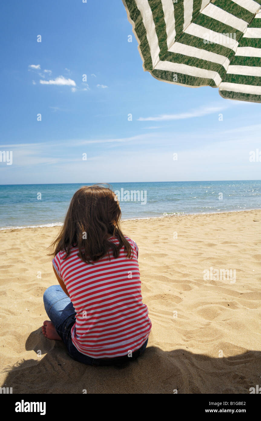 Teenage girl sitting on a sandy beach under a parasol in summertime. Stock Photo