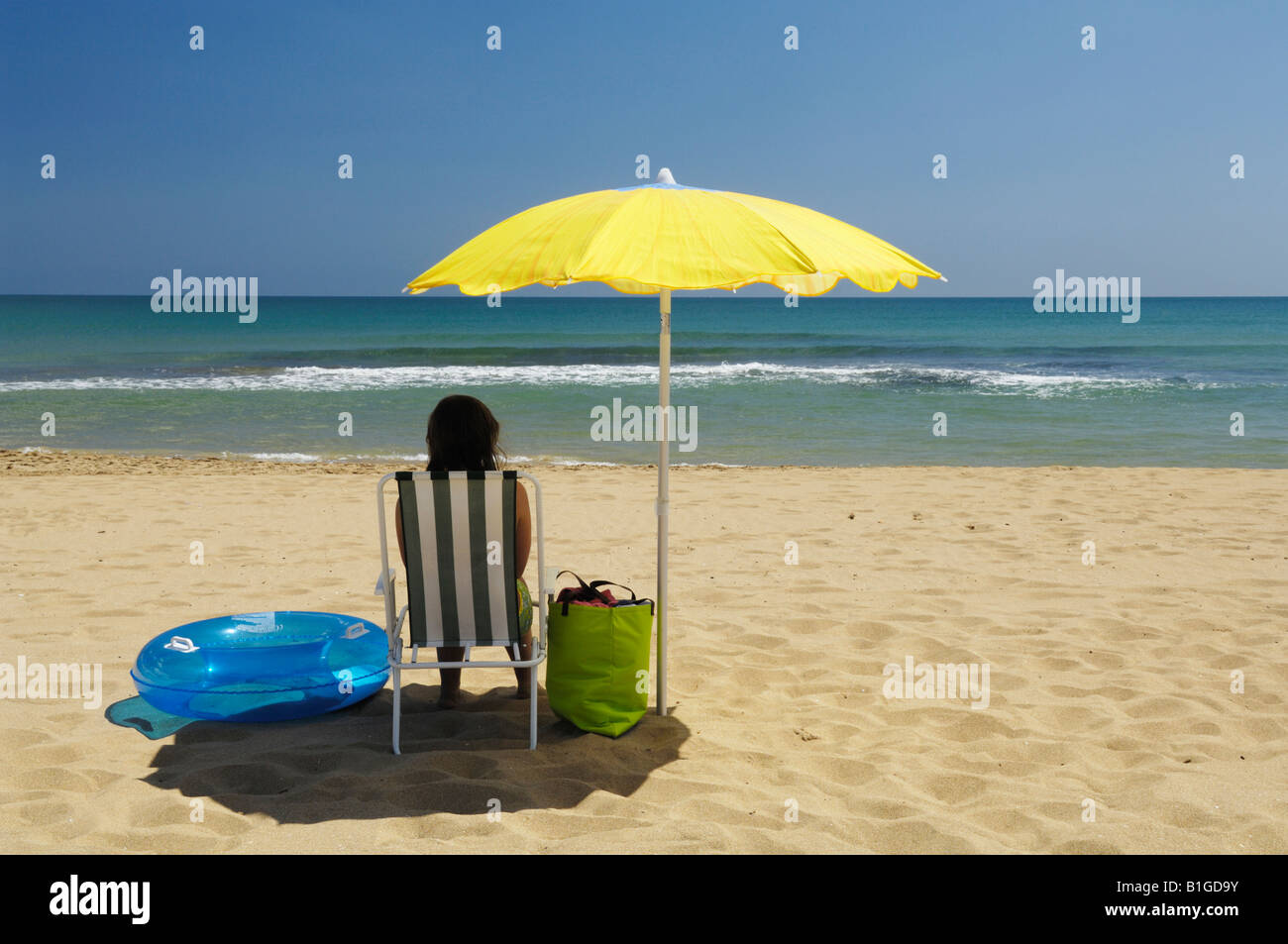 A teenage girl sitting under a parasol on a sandy beach by the sea in summertime. Stock Photo