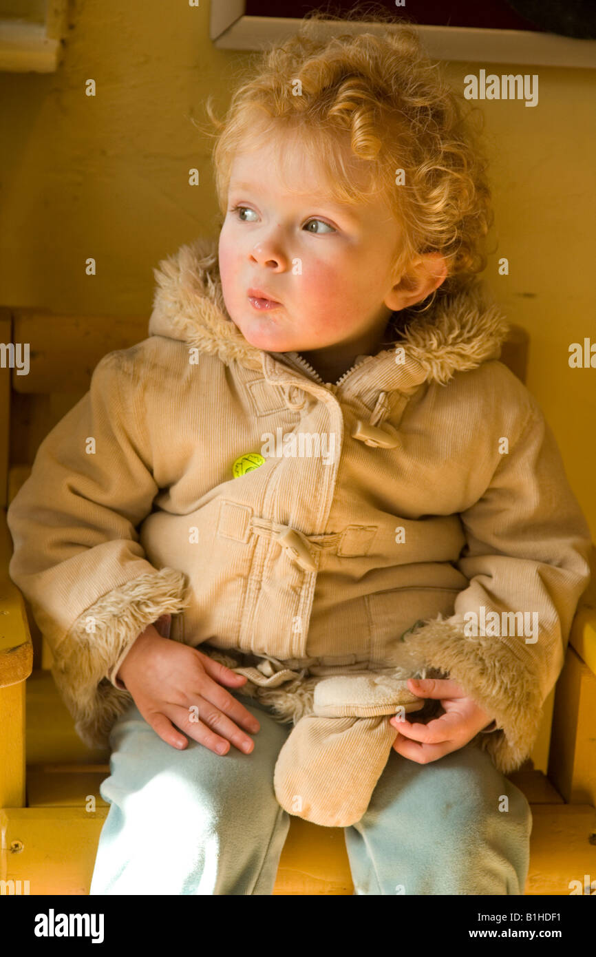 Strawberry blonde toddler wearing winter coat sitting on wooden seat Stock Photo