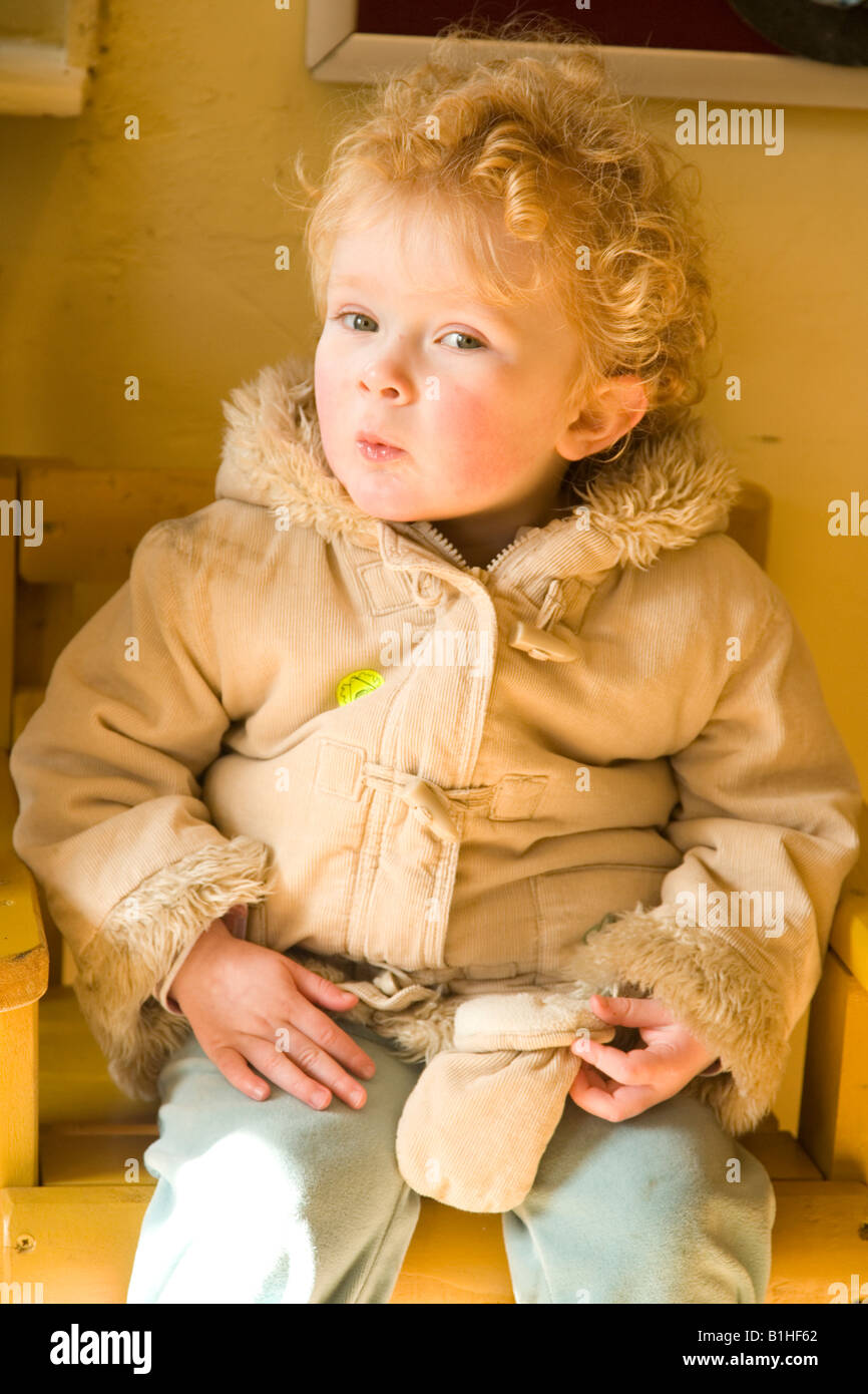 Strawberry blonde toddler wearing winter coat sitting on wooden seat Stock Photo