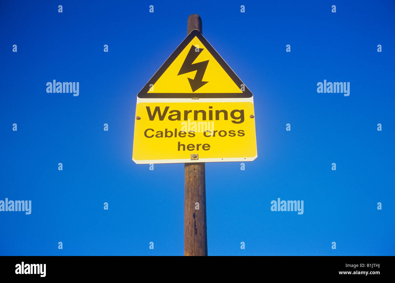 Electricity thunderbolt symbol in triangular yellow and black sign on pole with blue sky and words Warning Cables cross here Stock Photo