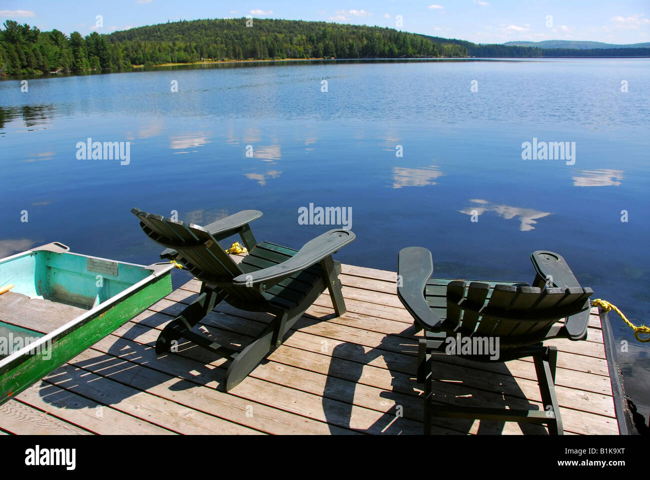 Two adirondack wooden chairs on dock facing a blue lake with clouds