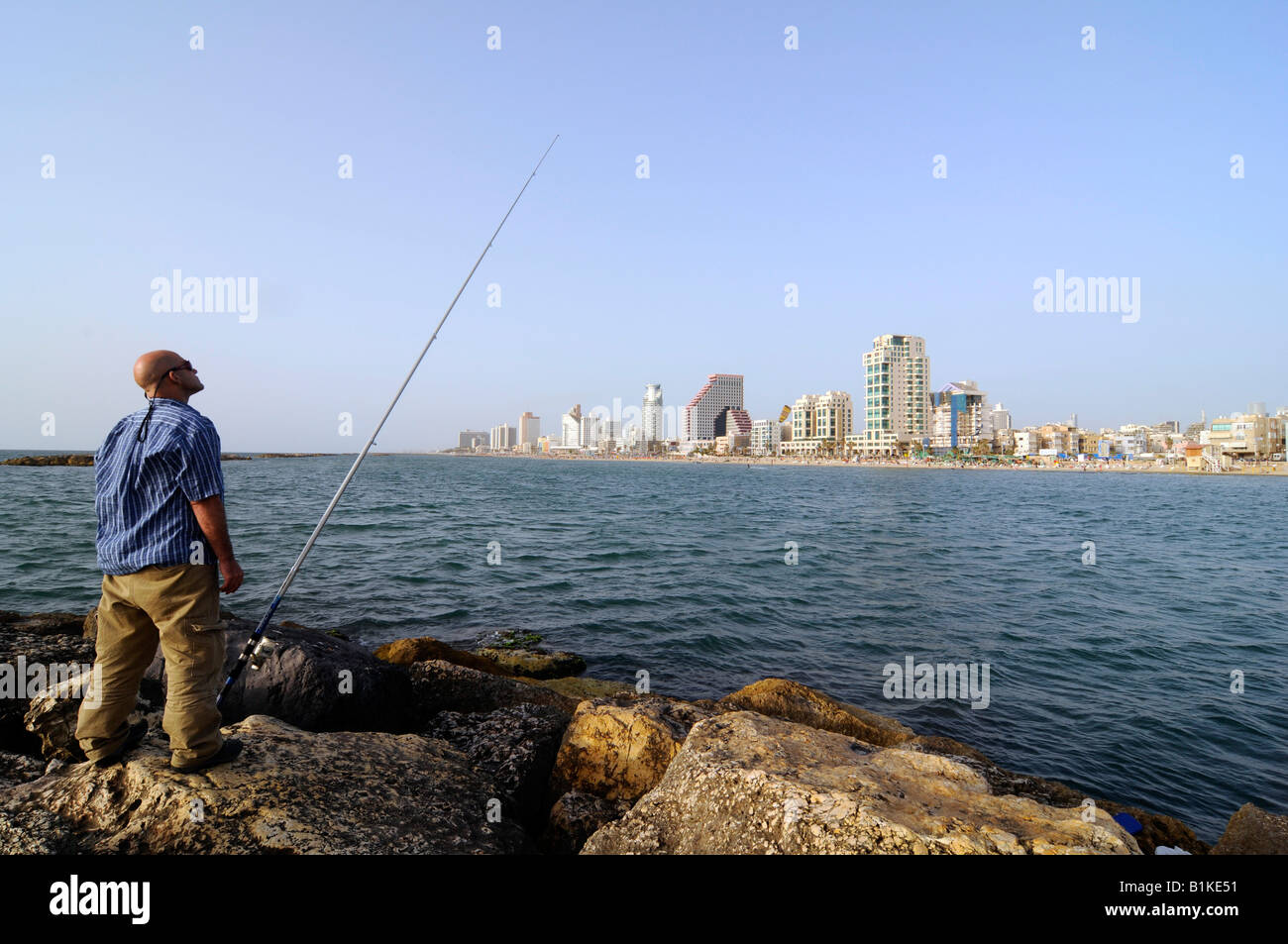 A man fishing near Tel Aviv beach, with the city's glittering skyscrapers skyline in background. Photo taken in Tel Aviv, Israel Stock Photo