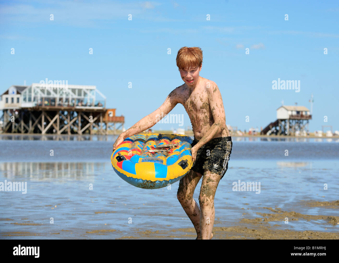 young boy covered in mud walking along a beach carrying an air mattress Stock Photo