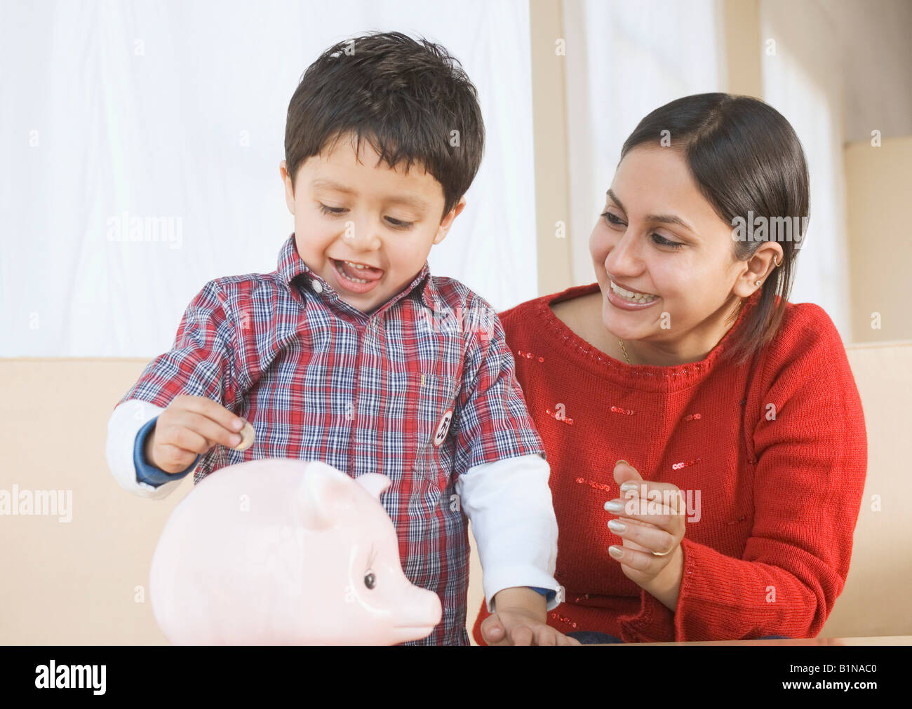 Boy putting a coin in a piggy bank with his mother sitting beside him ...