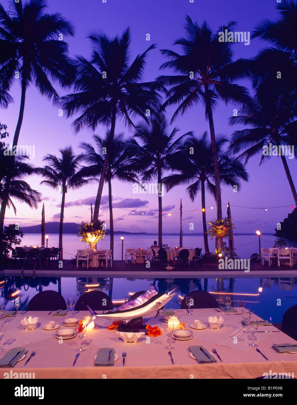 Elegant outdoor dining setup by a pool with palm trees and sunset in a tropical location Stock Photo