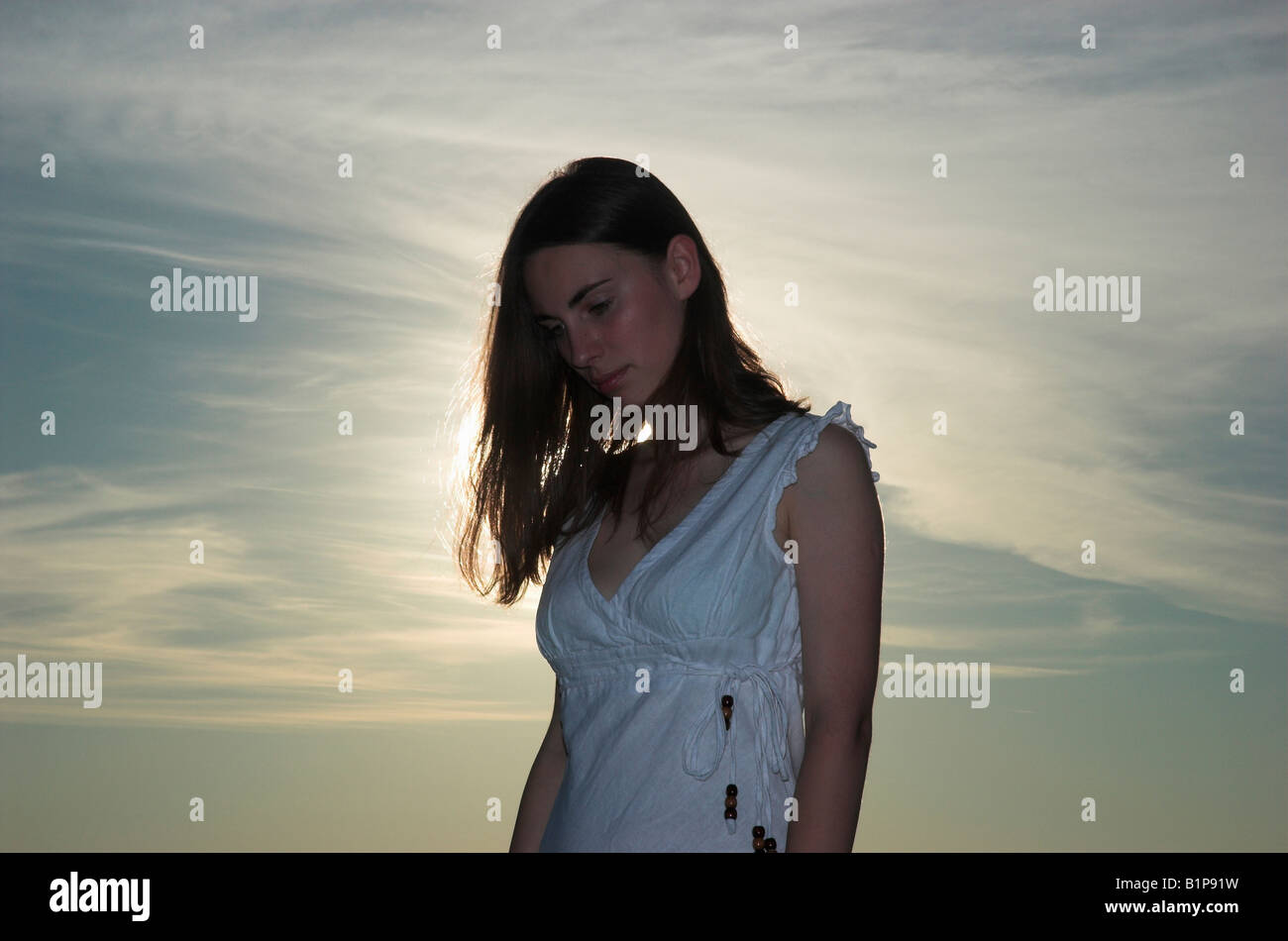 Silhouette of a young woman wearing white dress standing outdoors at dusk sun shining behind head bowed Stock Photo