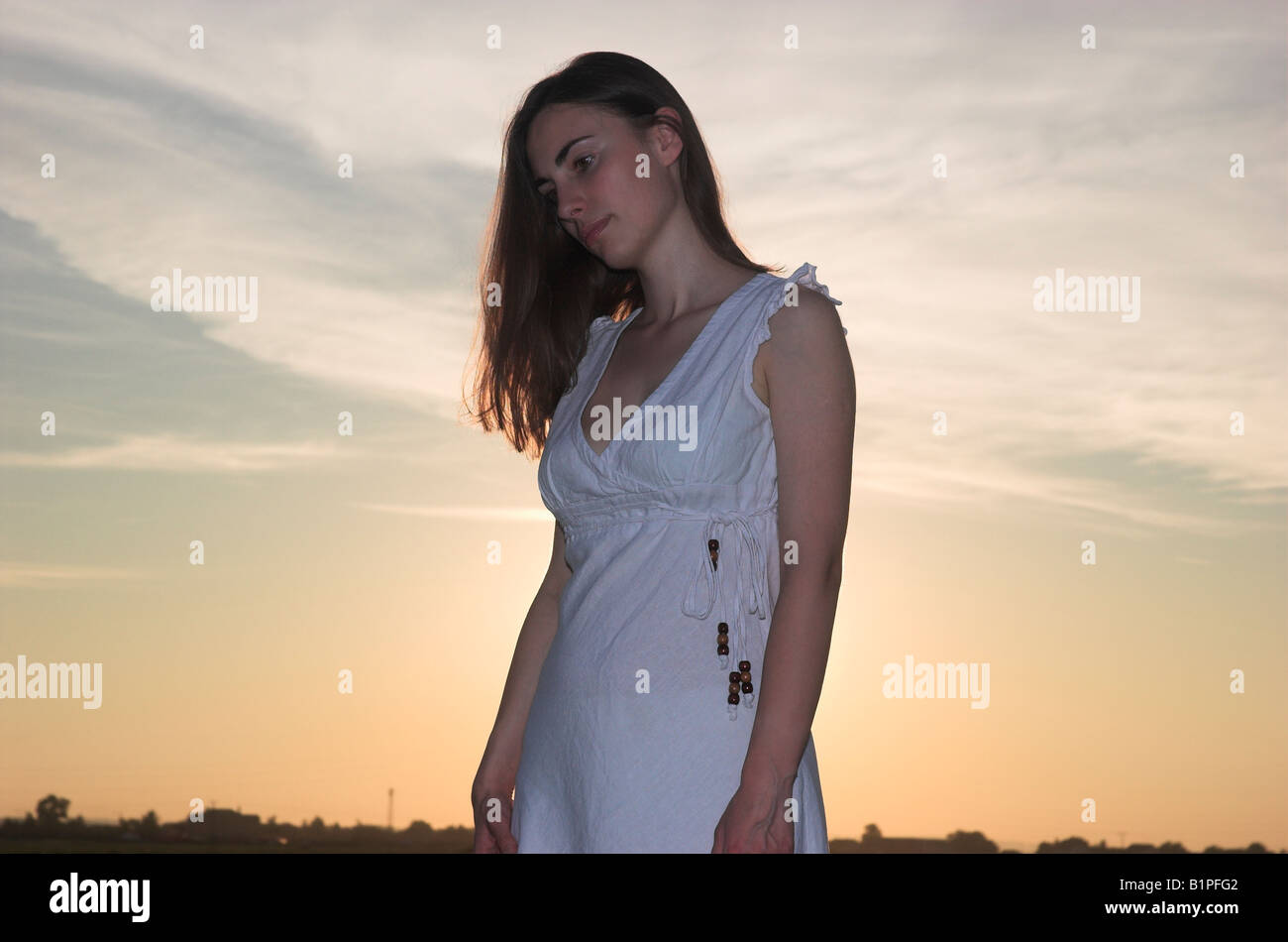 Silhouette of a young woman wearing white dress standing outdoors at dusk sun shining behind head bowed Stock Photo