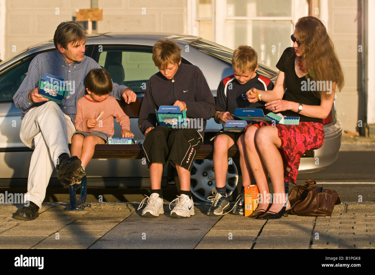 Family eating fish and chips convenience food sitting on a bench in sunshine mother father three sons, Aberystwyth Wales UK Stock Photo