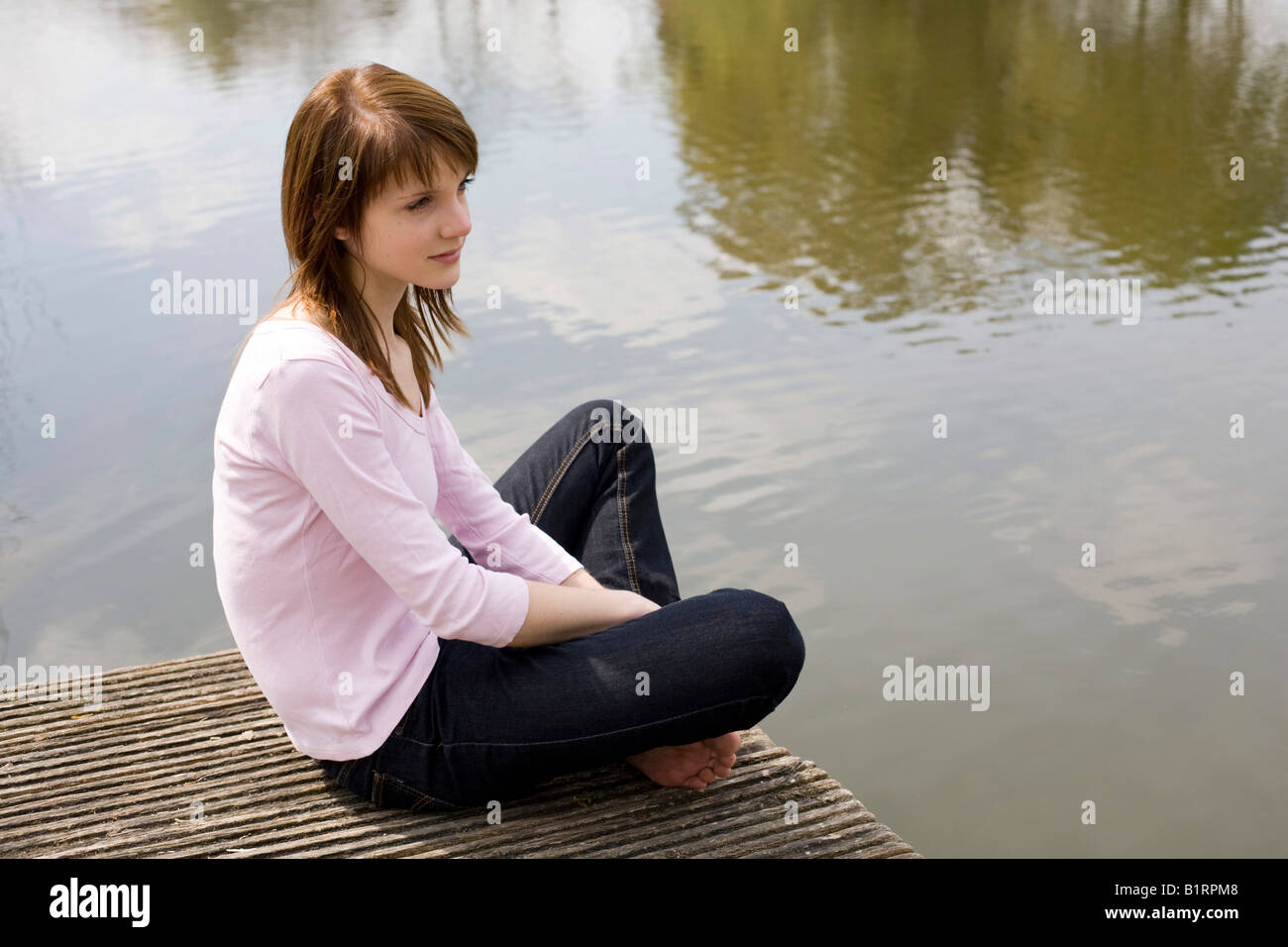 Young woman sitting on a jetty, wooden dock, looking at the water Stock Photo