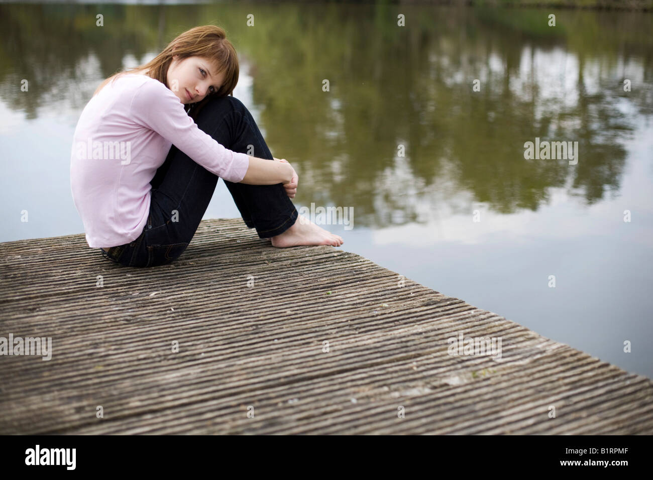 Young woman sitting on a jetty, wooden dock, looking pensive Stock Photo