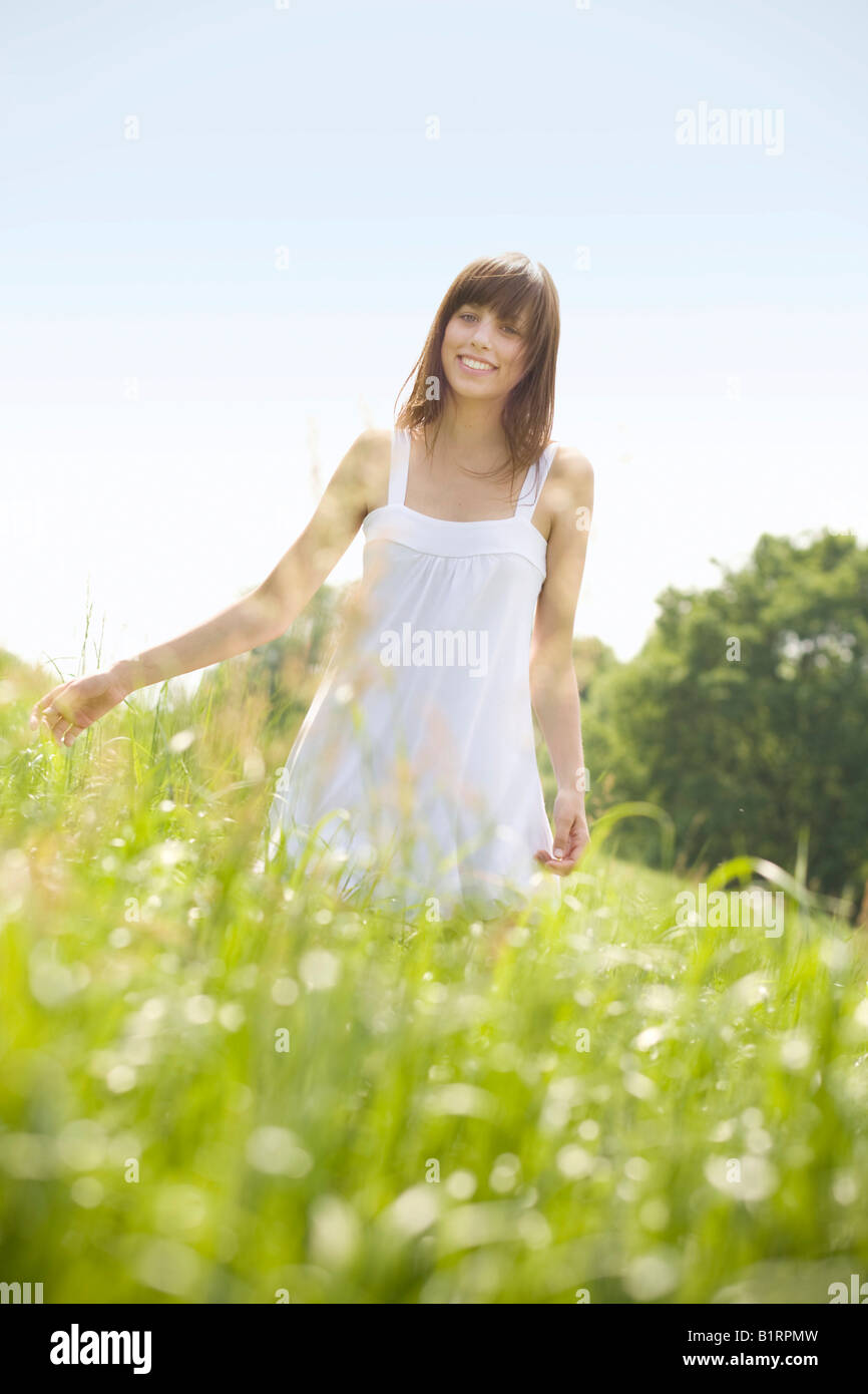 Young, dark-haired woman wearing a white dress standing in a summerly meadow, looking friendly at the camera Stock Photo