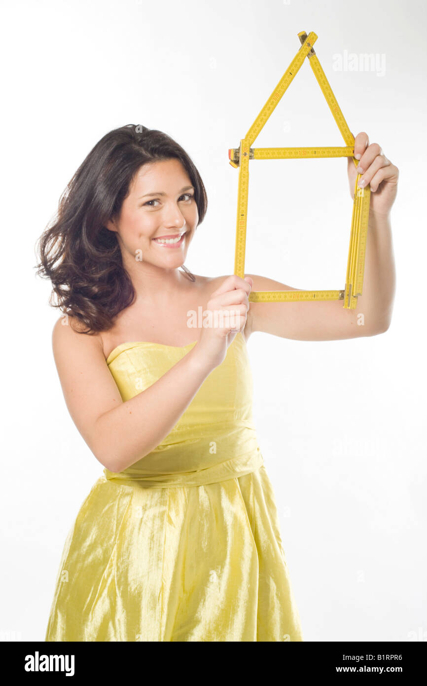 Young woman, wearing a yellow dress, smiling while holding in her hands a yellow folding ruler folded in the shape of a house Stock Photo