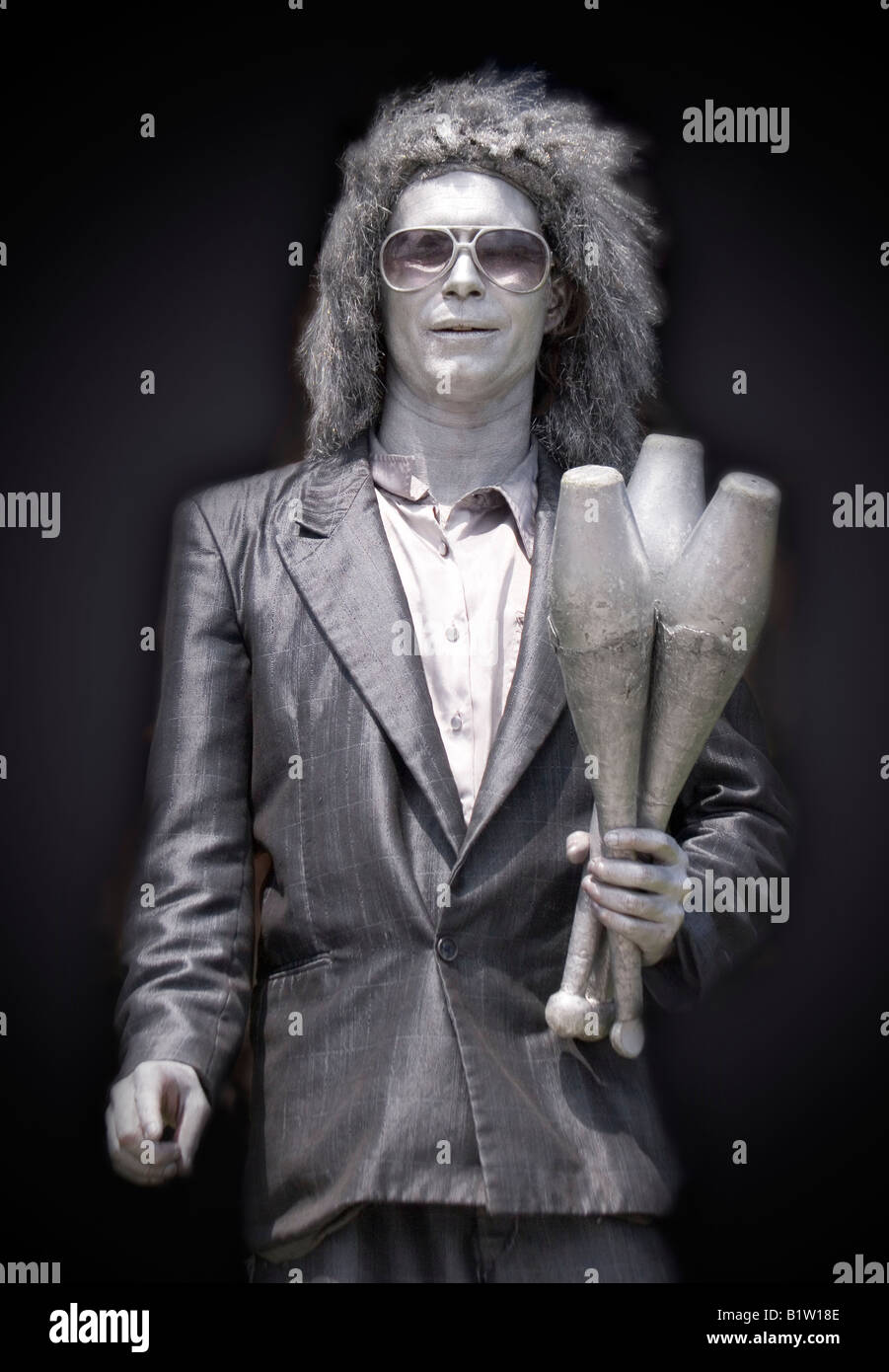 A street performer poses with his juggling clubs, dressed in silver clothing, wig and makeup. Stock Photo