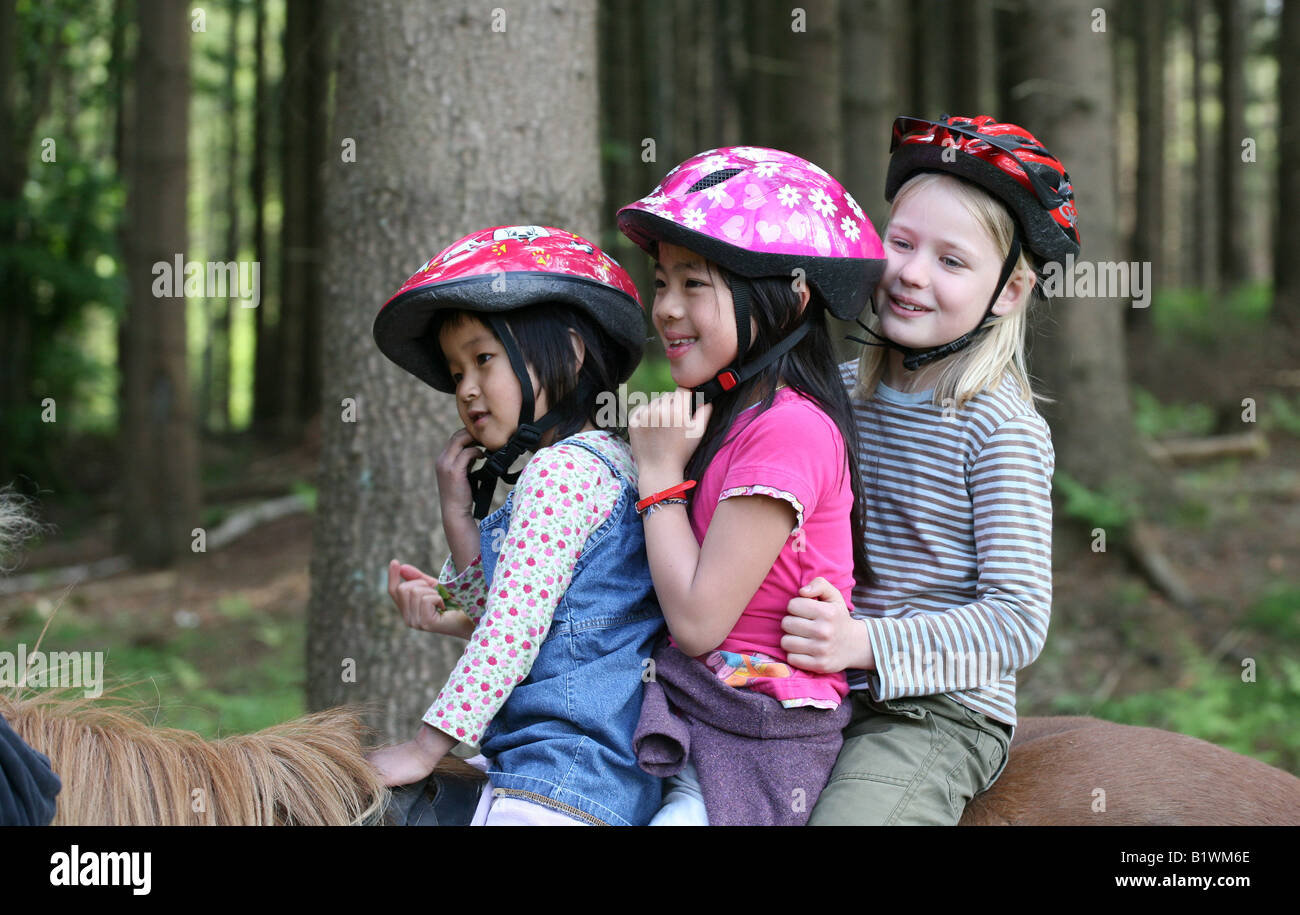 three friends one caucasian blond and two asian 9 years 5 years on horses in a forest Stock Photo