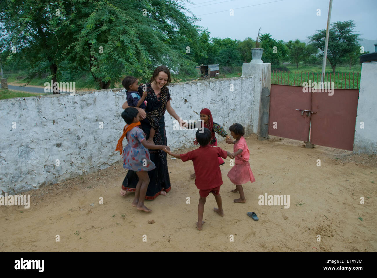 A young Western woman plays with local village children in the courtyard of a Rajasthani house, Thar desert Rajasthan, India. Stock Photo