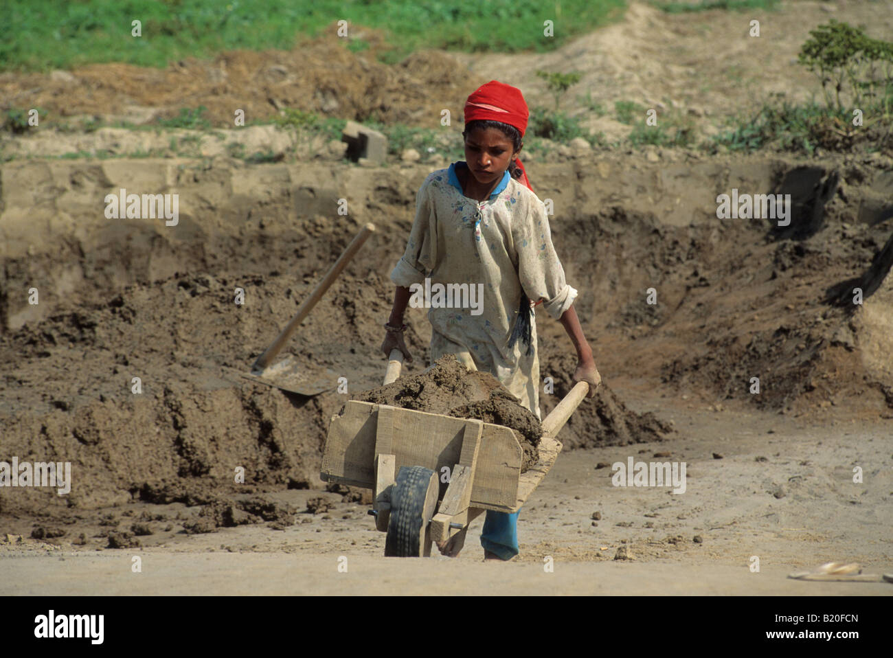 A young girl working as a child laborer in a brick kiln factory moves a wheelbarrow of dirt in Punjab Pakistan Stock Photo